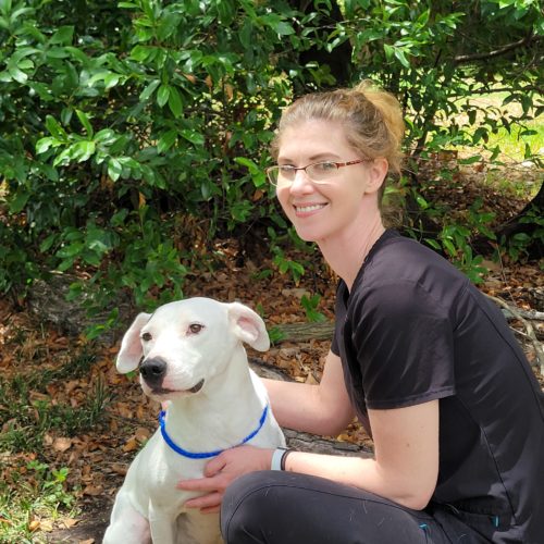 Britnee Doussan sitting outdoors with a white dog, both smiling, surrounded by green foliage.