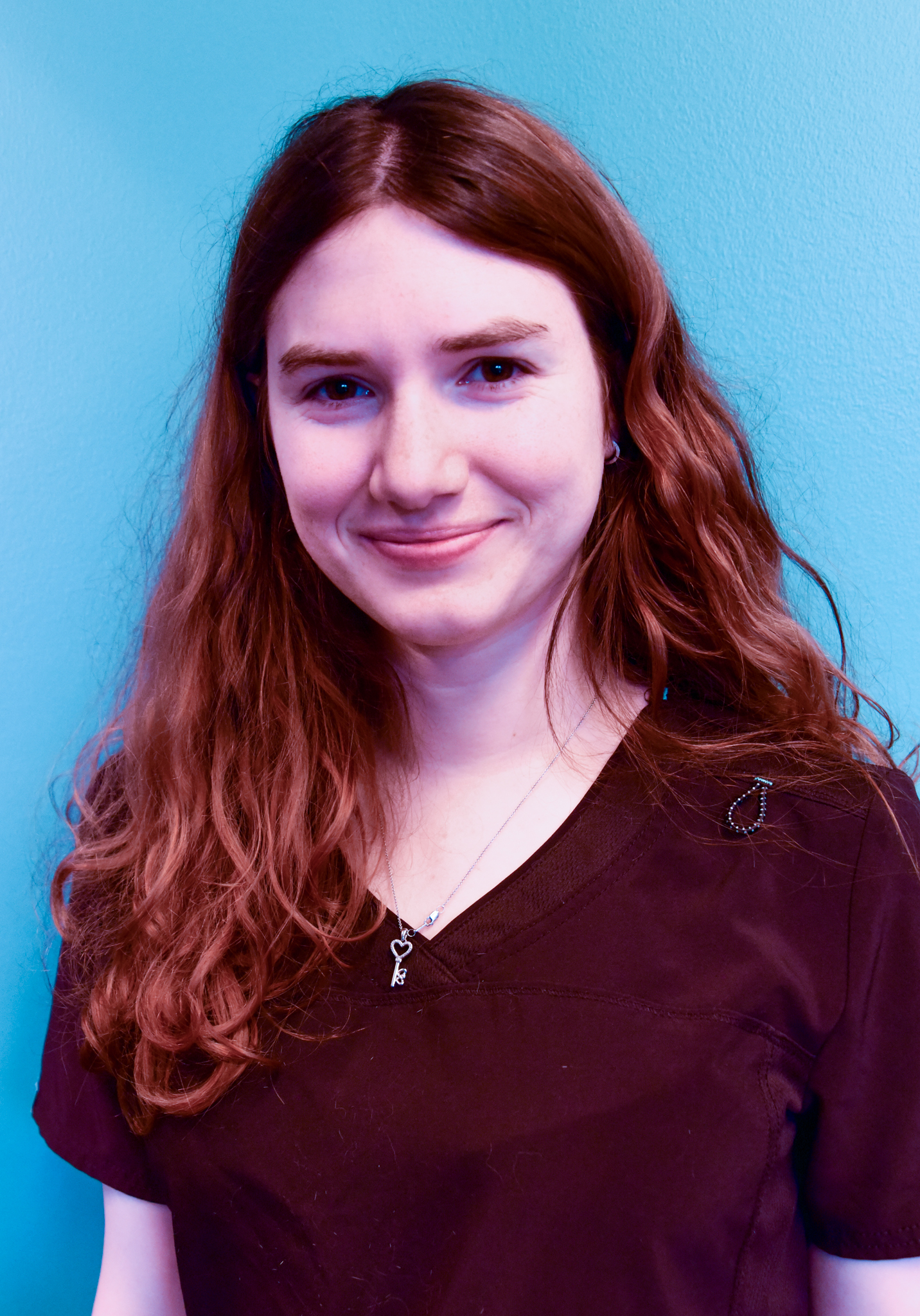 Young woman with long brown hair smiling against a teal background, wearing a dark scrub top.